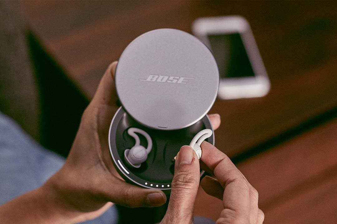 person holding sleepbuds in their charging case. background is an iphone displaying the Bose Sleep app.